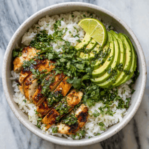 A bowl filled with white rice topped with sliced grilled chicken breast, fresh avocado slices, and chopped cilantro, all drizzled with a green cilantro-lime sauce. A lime wedge rests on the side, adding a bright pop of color. The dish is vibrant and fresh, served in a ceramic bowl on a marble surface.