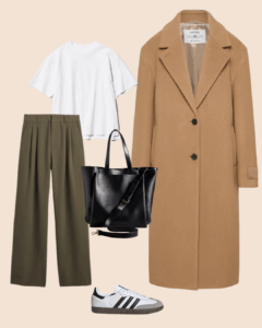 Flat lay of a minimalist fall outfit featuring a camel wool coat, white t-shirt, olive green wide-leg trousers, black leather tote bag, and white Adidas sneakers with black stripes on a beige background.